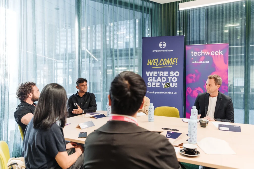 A diverse group of people sitting at a conference table engaged in discussion. Background features "Employment Hero" and "Techweek" banners, creating a welcoming atmosphere.