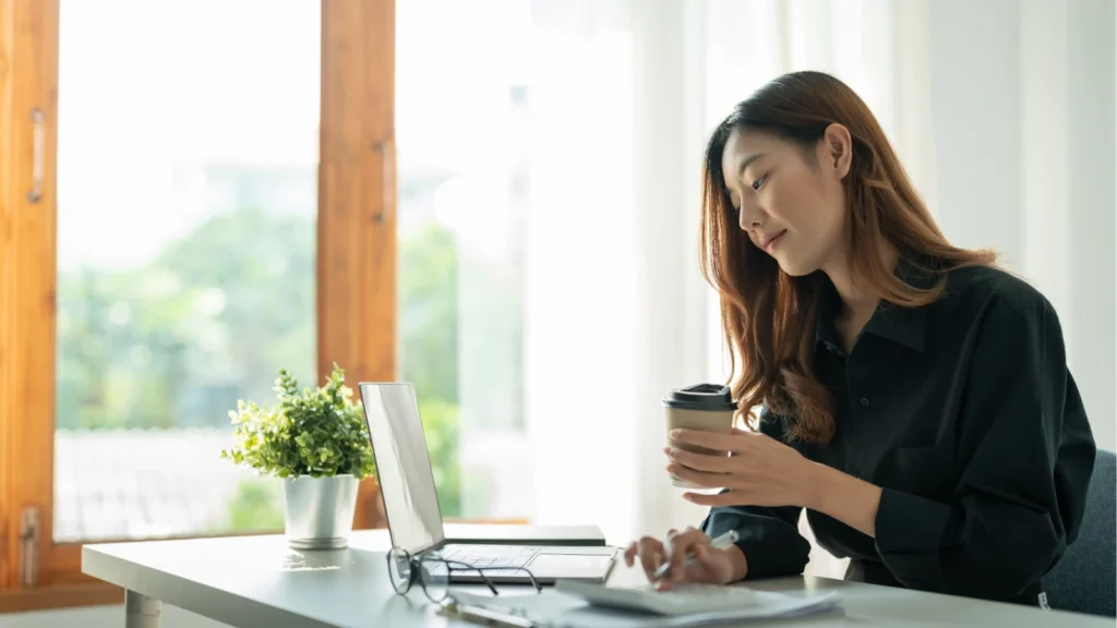 Asian businesswoman working at a laptop with a calculator and coffee cup at a bright desk, ideal for remote work concepts.