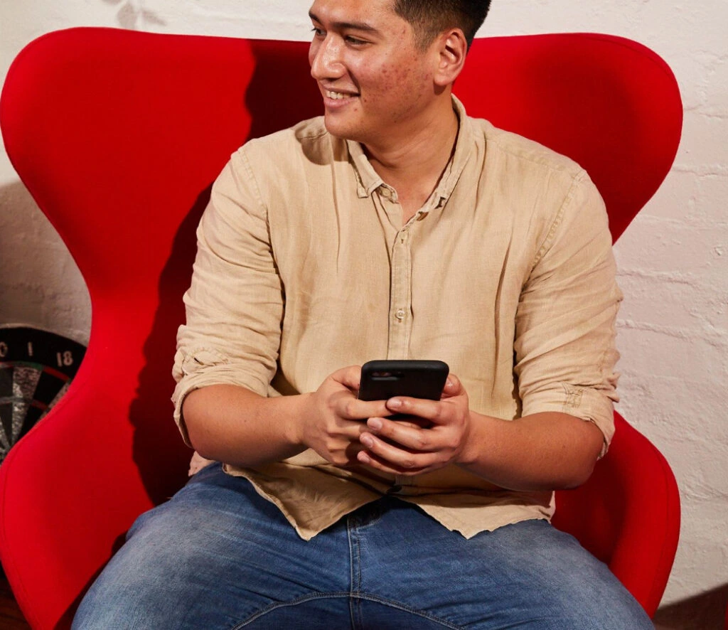 A person in a beige shirt and jeans, smiling and holding a phone, sits on a bright red, modern chair. The tone is casual and relaxed.