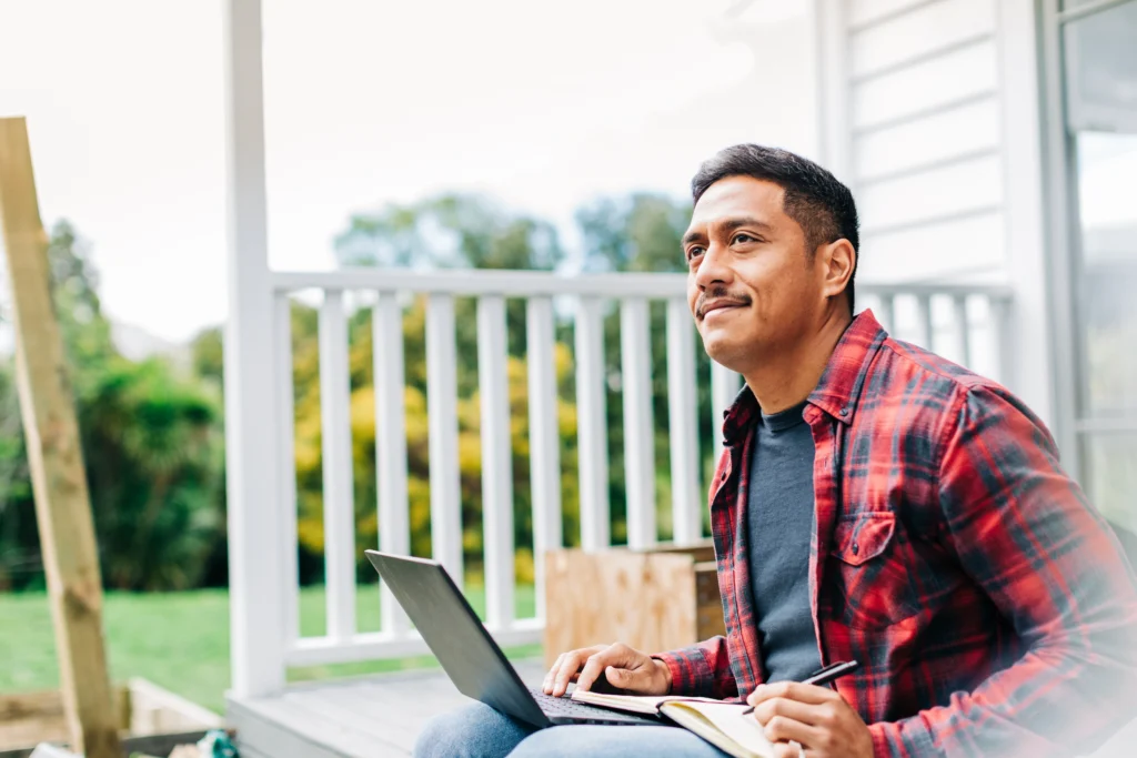 A man in a red plaid shirt sitting on a porch with a laptop and notebook, looking thoughtfully off-camera.