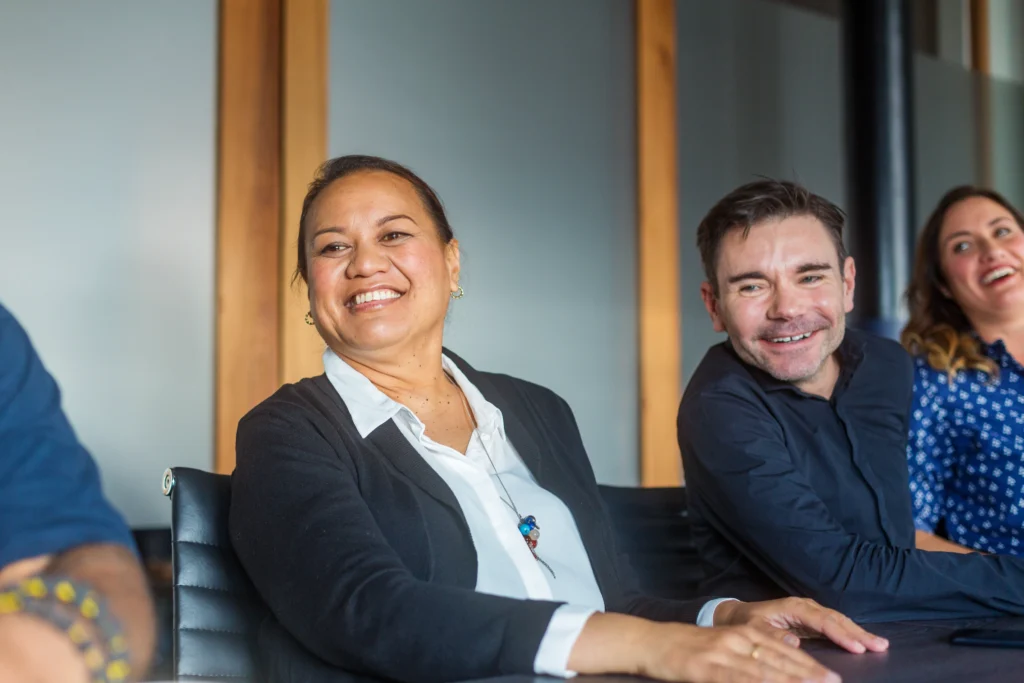 A group of three people sit at a conference table, smiling and laughing. The atmosphere is warm and friendly, suggesting a positive meeting or discussion.