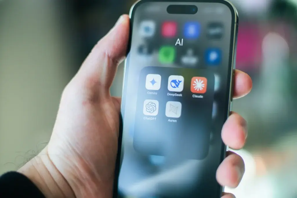A hand holding a smartphone showing an "AI" app folder containing Gemini, DeepSeek, Claude, ChatGPT, and Auren icons.