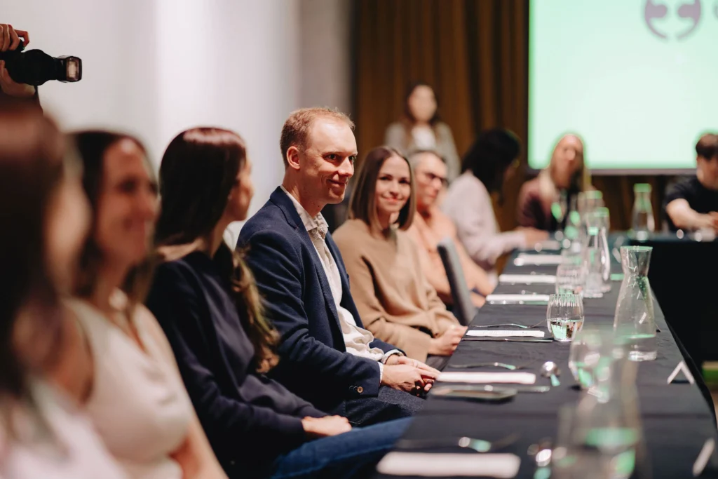 A group of people sit attentively at a long table set with water glasses and utensils, suggesting a formal meeting or conference. The atmosphere is focused and professional.