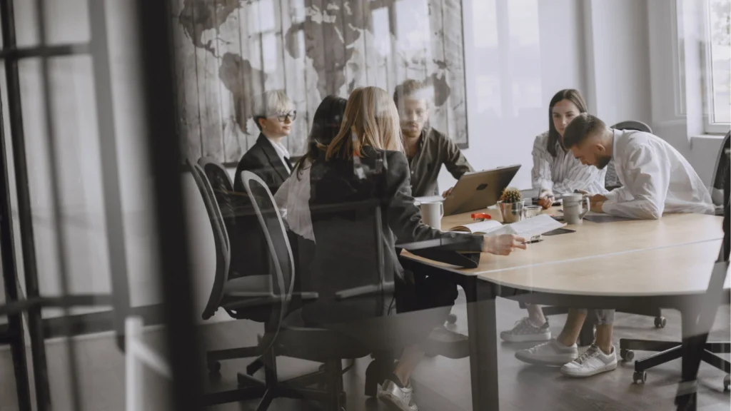 A diverse group of business professionals discussing documents and working on laptops around a large wooden table in a conference room.