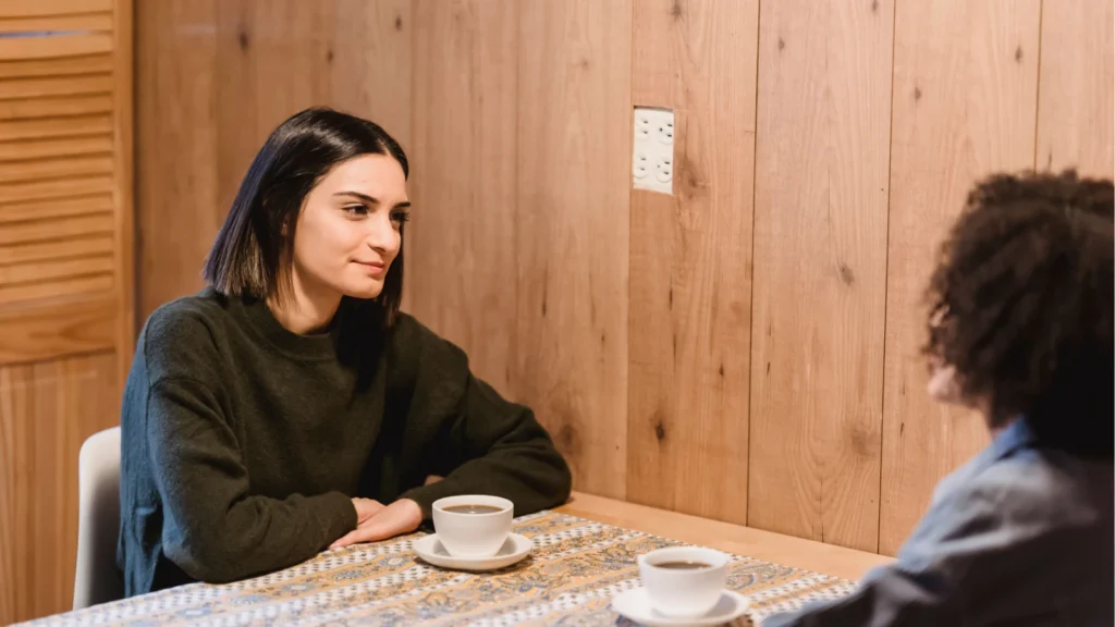 A young woman with dark hair sits at a wooden table with coffee cups, listening attentively to another person during a conversation.