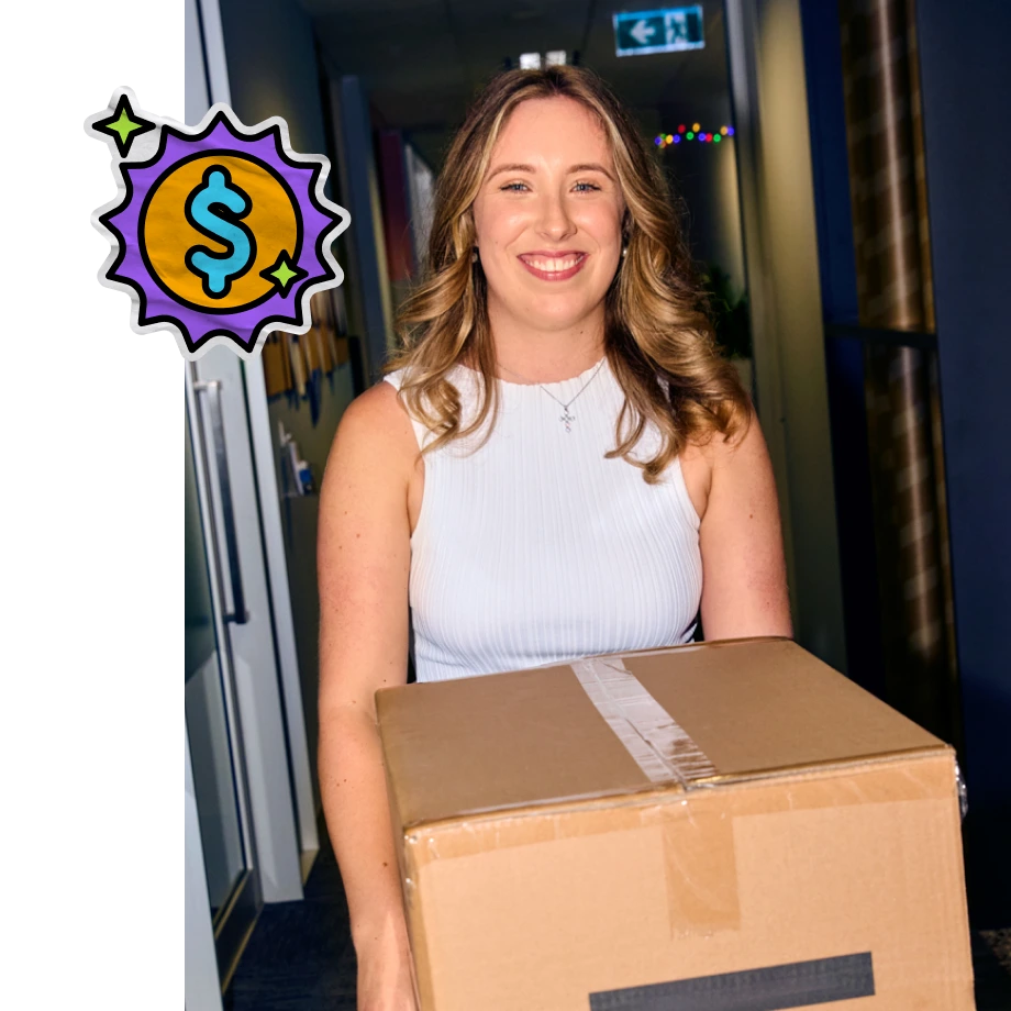 A smiling young woman with wavy brown hair wearing a white sleeveless top holds a large brown cardboard box in an office hallway, with a cartoonish graphic of a blue dollar sign inside a yellow and purple starburst overlaid in the top left corner.