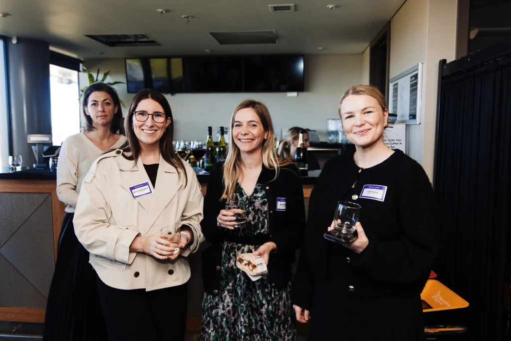 Four women hold drinks and smile at the camera while standing near the bar area during a corporate networking event.
