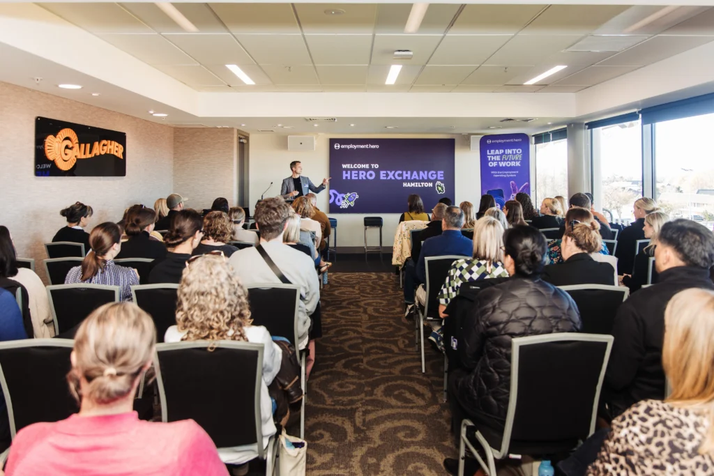 An audience seated in a conference room listens to a speaker presenting onstage at the Hero Exchange Hamilton event.
