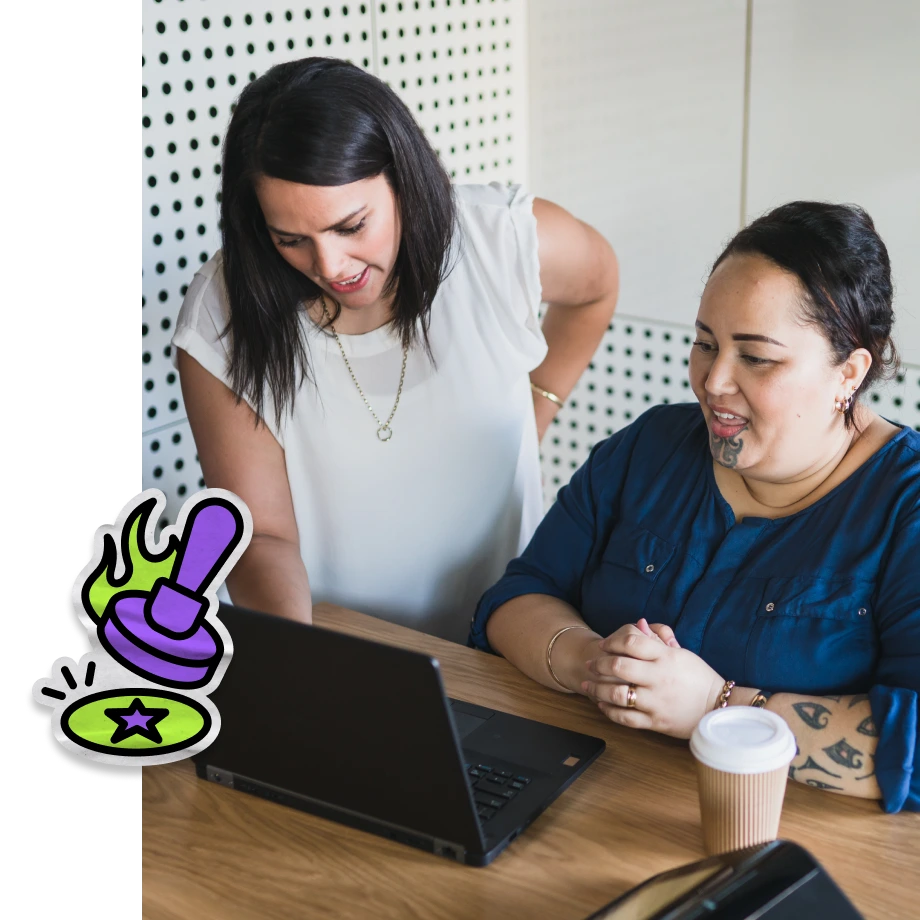 Two women are looking at a black laptop on a wooden desk in front of a white pegboard wall. The woman on the left, wearing a white sleeveless top, leans over to look at the screen, while the seated woman on the right, wearing a blue shirt, has a facial tattoo on her chin (moko kauae) and tattoos on her arm. A paper coffee cup sits on the desk, and a cartoonish graphic of a purple stamp pressing onto a green star circle is overlaid in the bottom left corner.