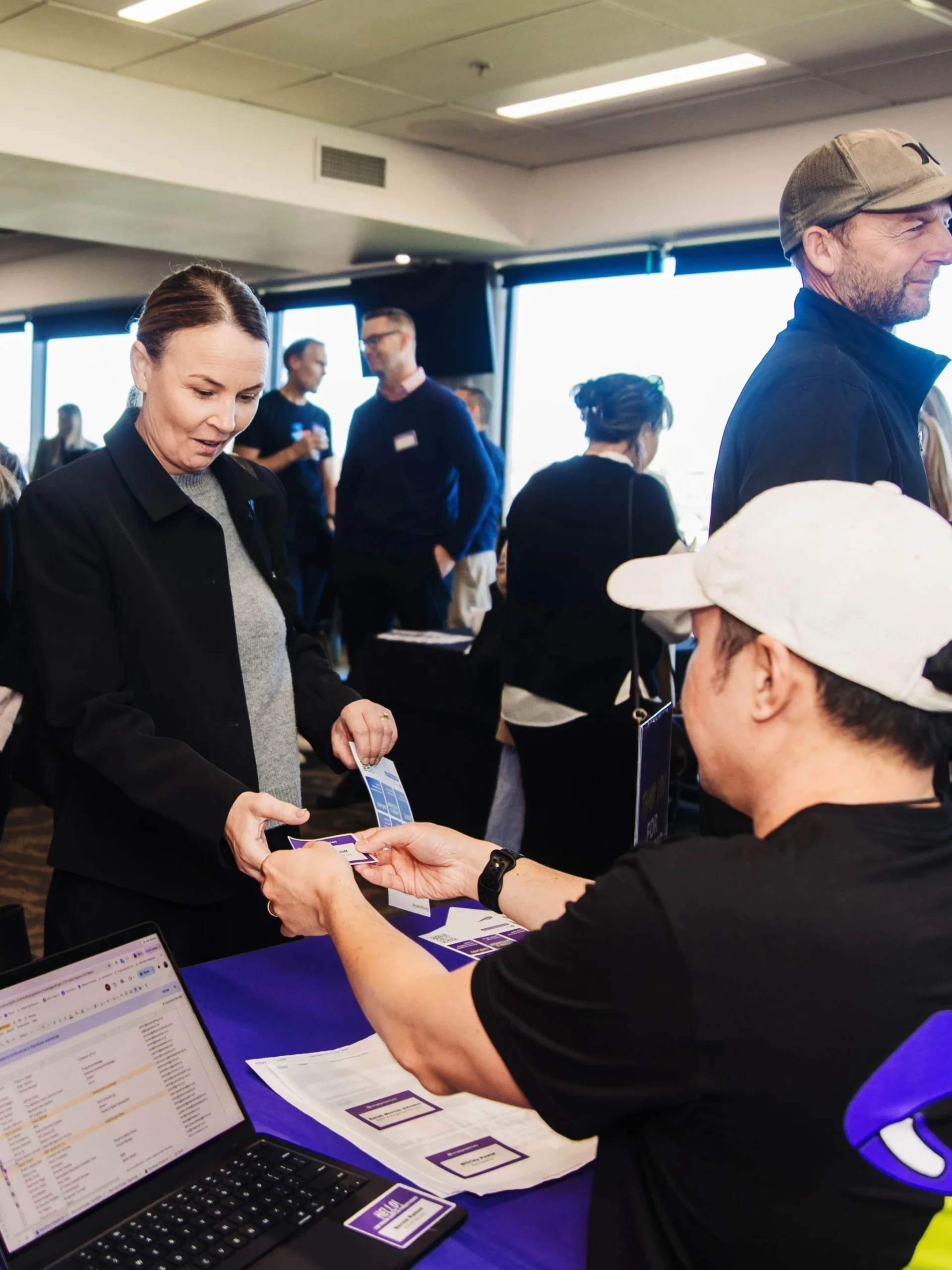 People are gathered at an event registration table covered with a purple cloth and a "REGISTER HERE" sign. A man wearing a white cap and black t-shirt sits behind the desk with an open laptop, handing a card to a woman in a black blazer, while another woman in a bright pink sweater and blue shoulder bag stands beside her. Several other attendees are mingling in the background.
