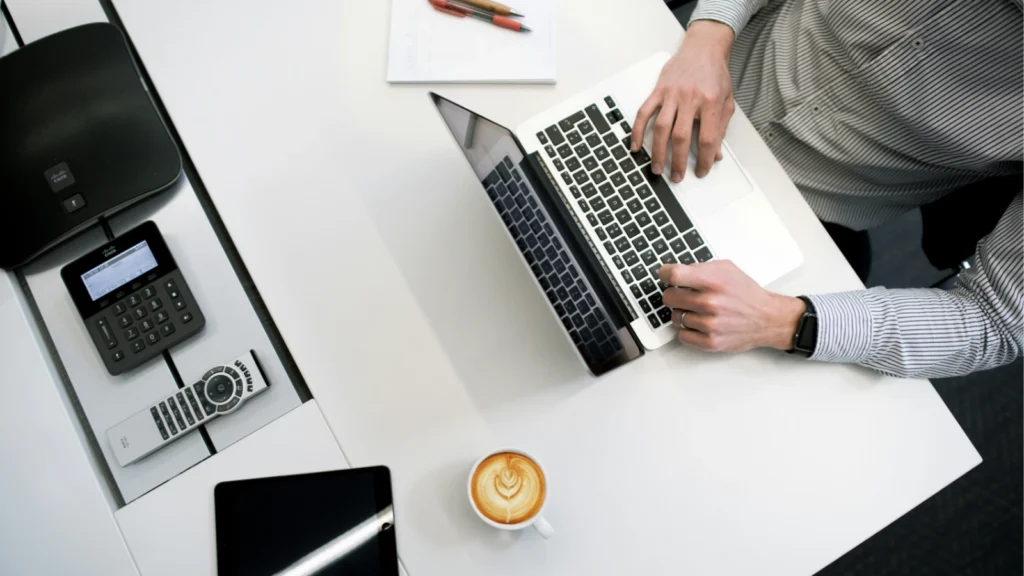 An overhead view of a person typing on a laptop at a neat white office desk alongside a cup of latte art and tech accessories.
