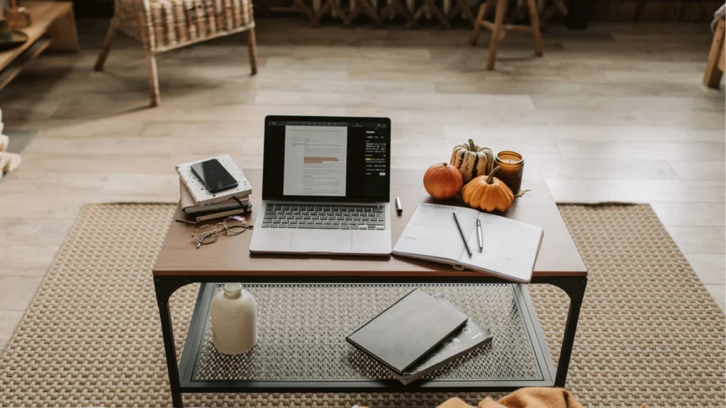 An open laptop sits on a coffee table alongside notebooks, decorative autumn pumpkins, glasses, and a candle.