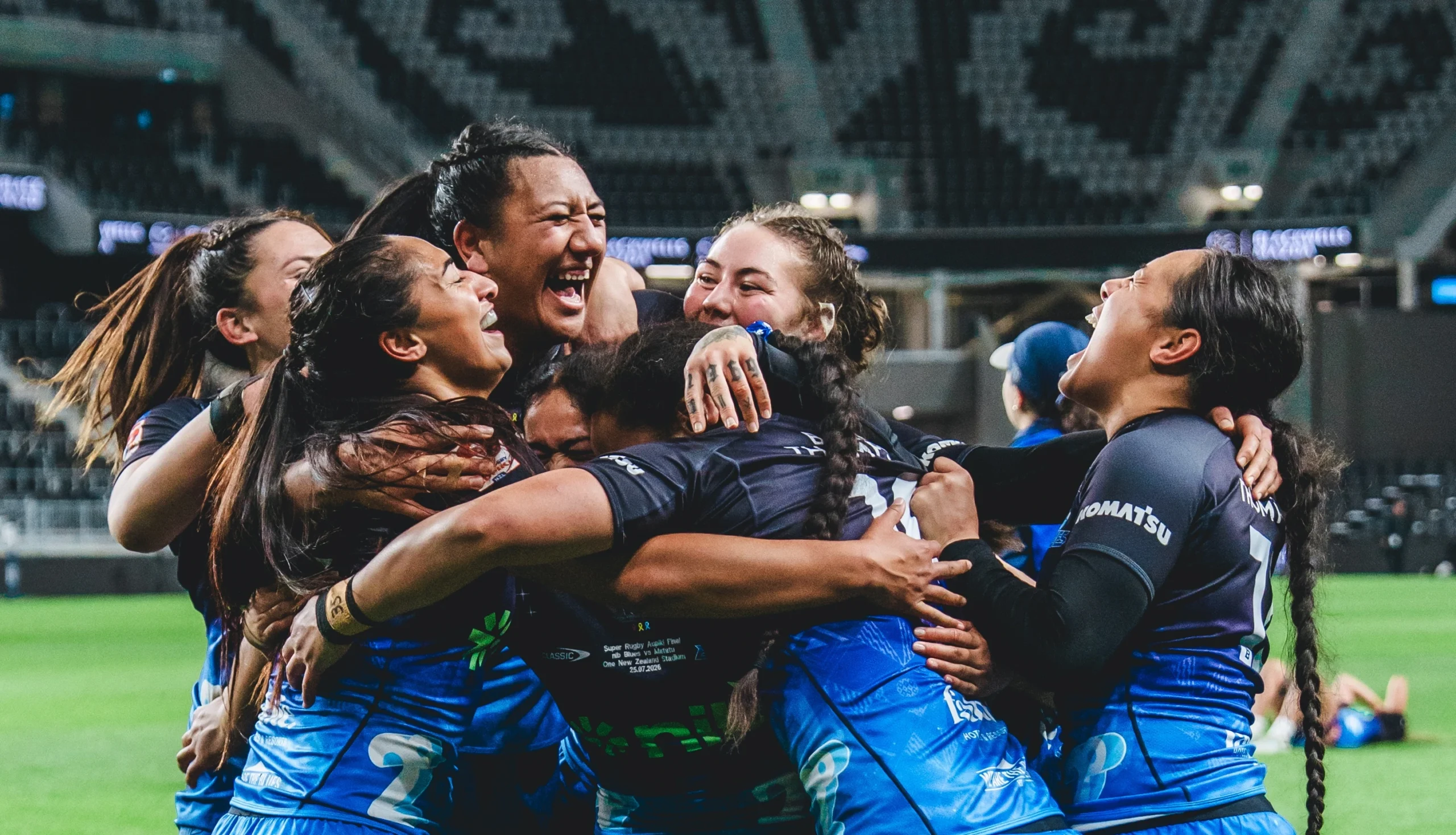 A team of female rugby players in blue jerseys huddle and celebrate joyfully on the field inside a large stadium.