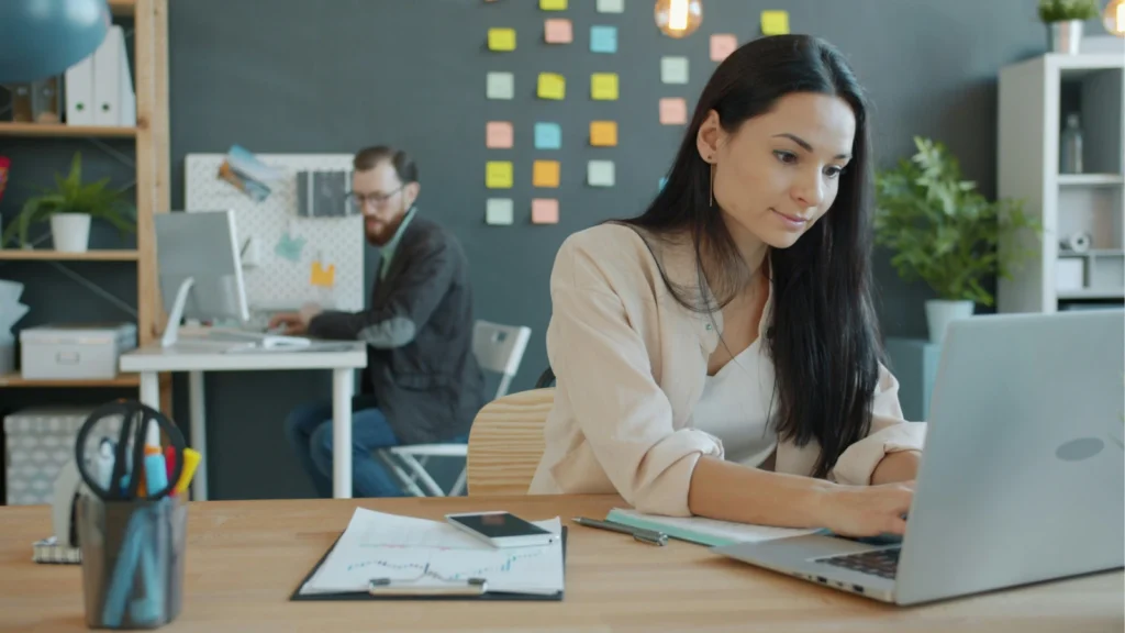 A focused woman works on her laptop at a wooden desk while a male colleague works at a computer in the background office space.
