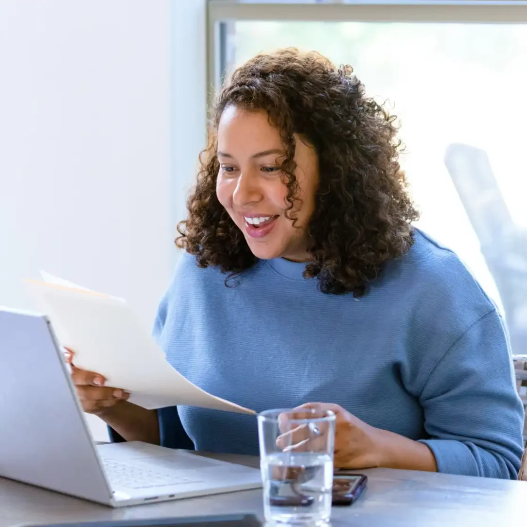 A bright, lifestyle photograph of a smiling woman with curly hair, wearing a light blue sweater. She is seated at a table, holding a paper document in one hand while happily looking at a silver laptop screen. A glass of water and a smartphone rest on the table beside her.
