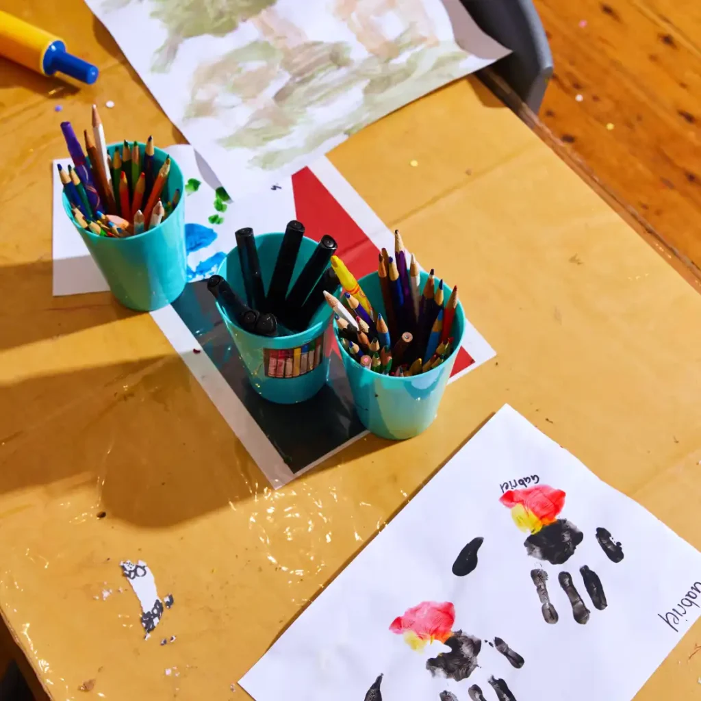 Here is the latest addition to our digital asset library:

Childcare_Hero@2x-1024x1024.webp: A close-up overhead shot of a wooden art table in a childcare or early learning setting. Three bright teal cups hold colored pencils and black markers, resting among scattered pieces of children's artwork. In the foreground, a white sheet of paper displays colorful painted handprints in black, red, and yellow, signed with the name "Gabriel." A yellow and blue toy rolling pin is visible in the top left corner.