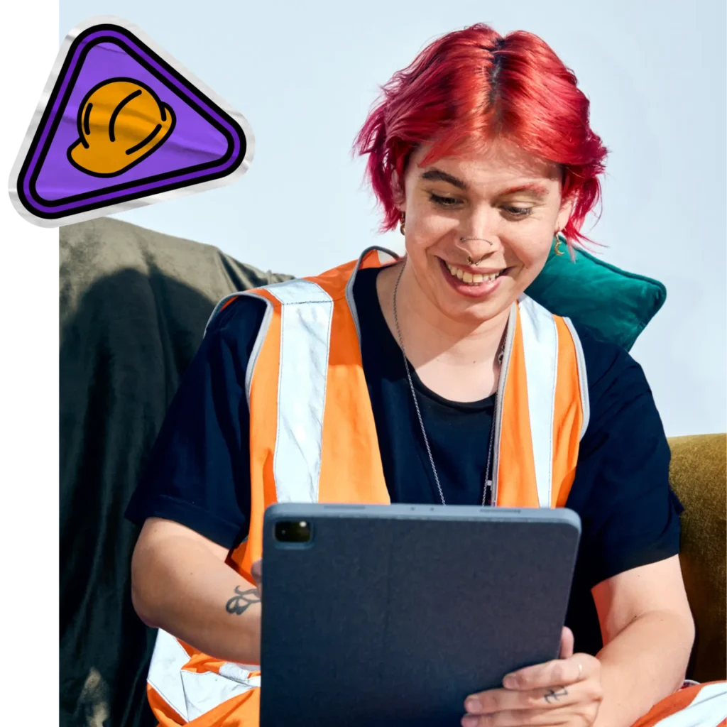 A smiling individual with bright red hair and facial piercings is seated outdoors, wearing a black t-shirt and an orange high-visibility construction vest. They are looking down happily at a grey tablet they are holding. In the top left corner, there is a branded cartoon graphic overlay featuring a yellow hard hat inside a rounded purple triangle.