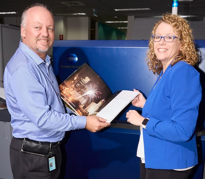 Two smiling colleagues hold open a printed photo booklet in front of a large blue printing machine.