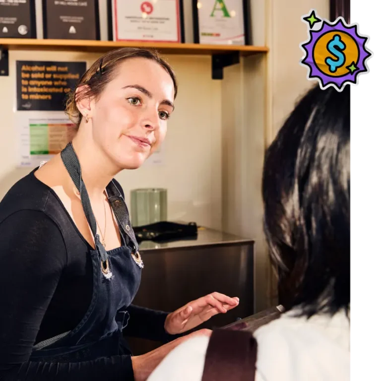 A young female employee wearing a dark denim apron over a black long-sleeved shirt stands behind a counter, attentively looking at a customer whose back is to the camera. The setting appears to be a cafe or retail shop, with framed certificates and an alcohol service policy sign visible on the wall behind her. A cartoonish graphic featuring a blue dollar sign inside a yellow and purple starburst is overlaid in the top right corner.