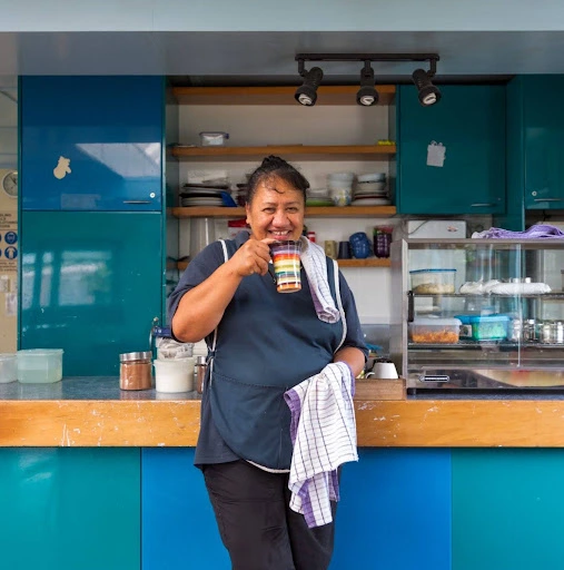 Smiling café worker holds a colorful drink and towel behind a wooden counter, framed by turquoise cabinets and shelves of dishes.