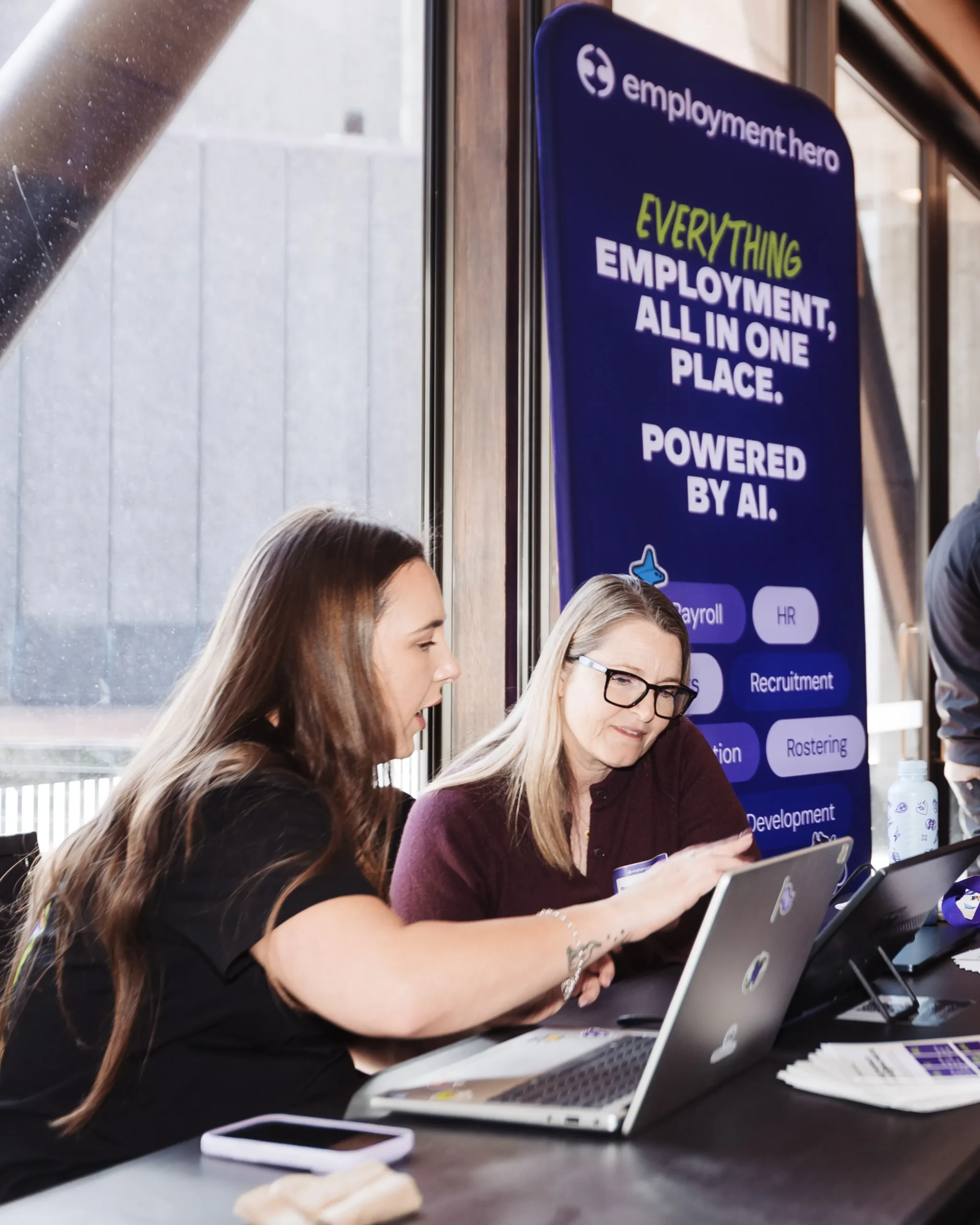 Two women work at a check-in desk with laptops next to an Employment Hero banner, while attendees mingle in the background.