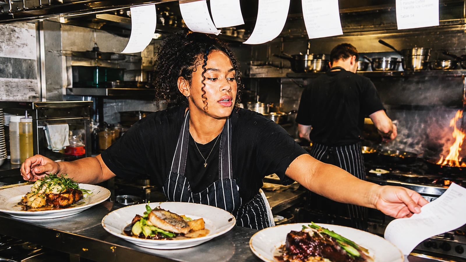 A chef in a striped apron reaches for an order ticket at a busy restaurant pass filled with plated meals.