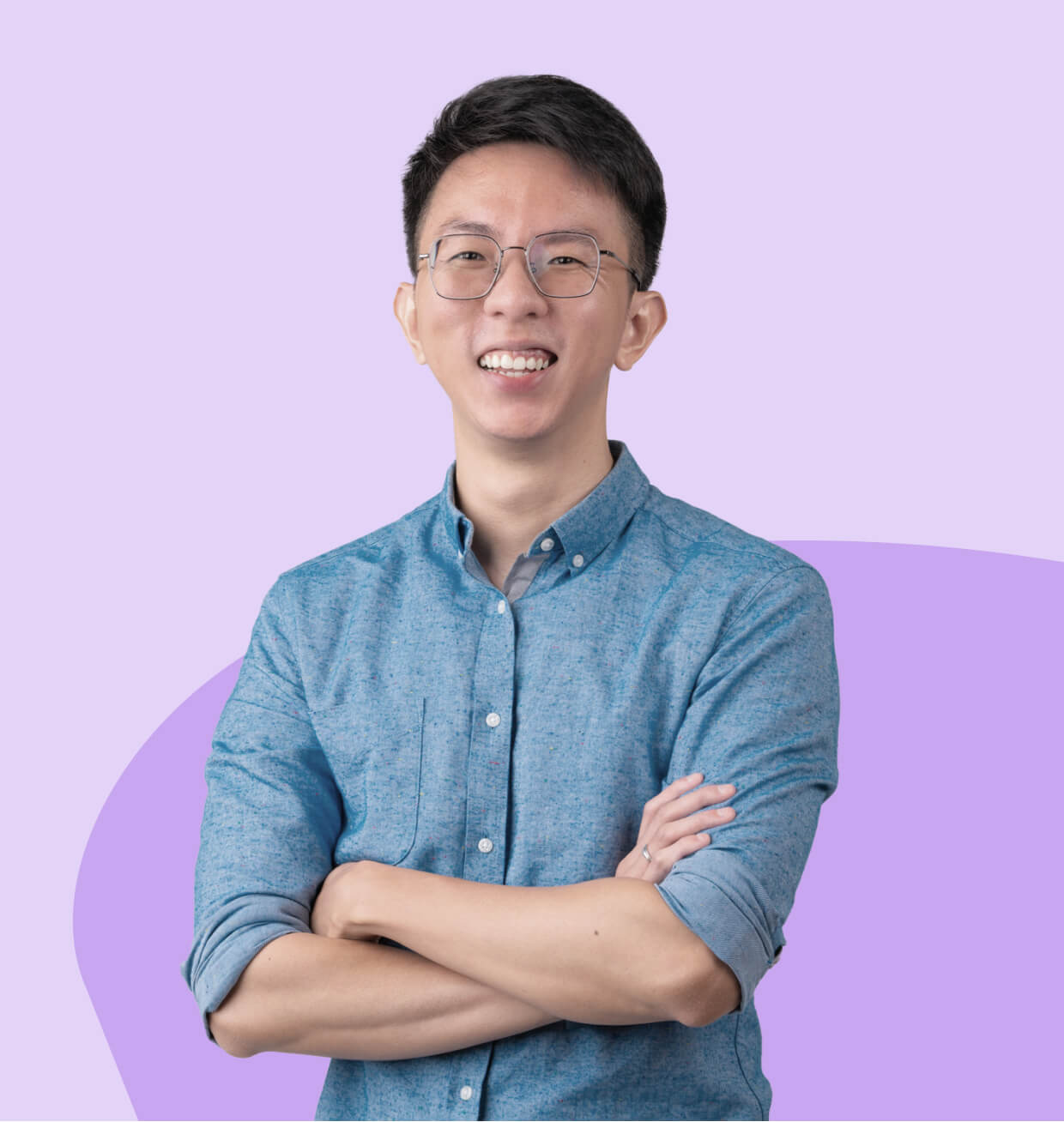 A smiling man wearing glasses and a blue button-down shirt, standing with arms crossed against a light purple background with abstract shapes.