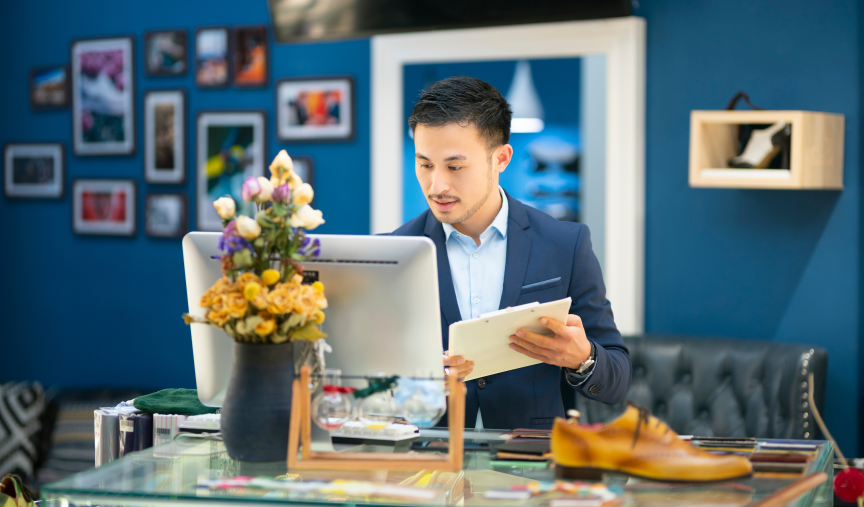 Man in a suit working at a retail store counter, looking at a computer screen while holding a clipboard. The counter has a floral arrangement and a shoe on display. The background features a blue wall with framed photos.