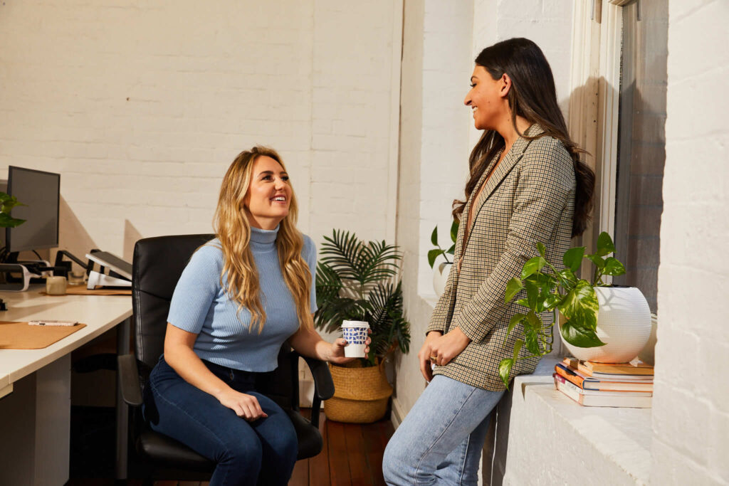 two women talking in the office, with one leaning on the wall and the other sitting on an office chair