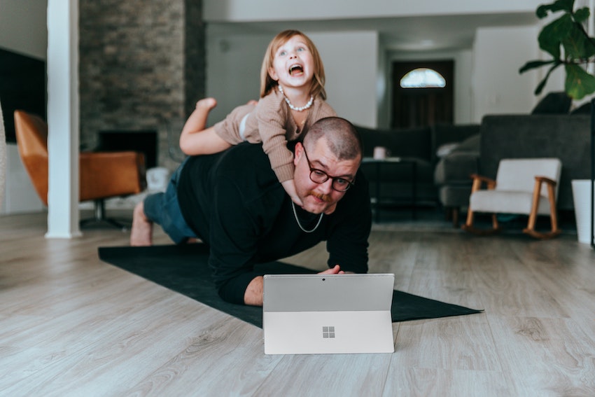 A father in plank position on a yoga mat looking at a tablet with a young girl on his back