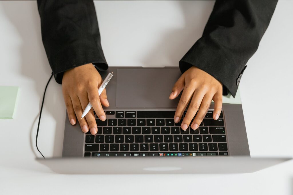 Close-up of a person in a black blazer using a laptop, holding a pen in one hand while typing with the other on a white desk.