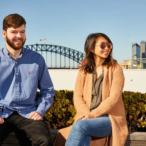 Two people sit outdoors, smiling, with the Sydney Harbour Bridge in the background. It's a sunny day, conveying a relaxed, joyful atmosphere.