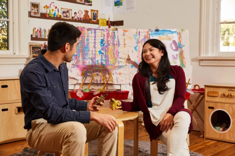 A man and woman sit at a small table in a colorful playroom, smiling and talking. Art supplies and children's paintings are visible in the background.