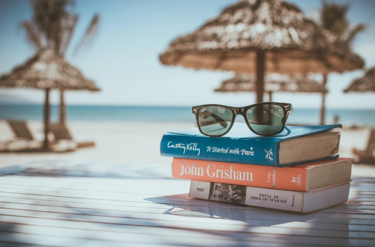 Sunglasses rest on a stack of books on a sunlit beach table. In the background, straw umbrellas and the ocean create a relaxed, vacation vibe.