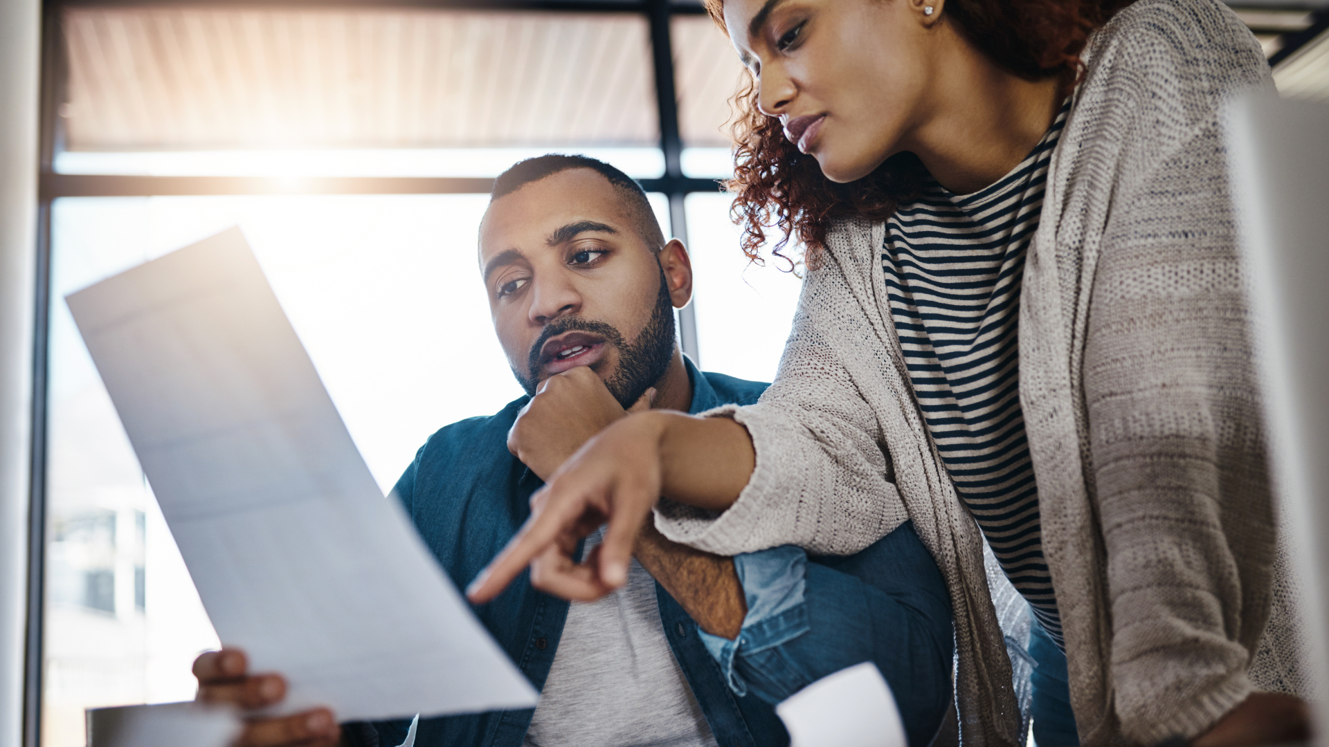 A man and woman are reviewing a document together. The man, seated, looks intently at the paper while the woman stands beside him, pointing at the document, seemingly explaining or highlighting important information. They appear to be in a well-lit office environment.