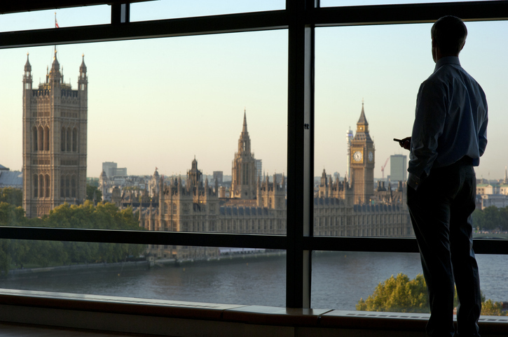 Silhouette of a person looking out a large window with a view of the iconic British Parliament buildings, including Big Ben, across the River Thames at dusk.