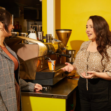 Two women talking near a coffee machine in a café, one gesturing while speaking and the other listening.