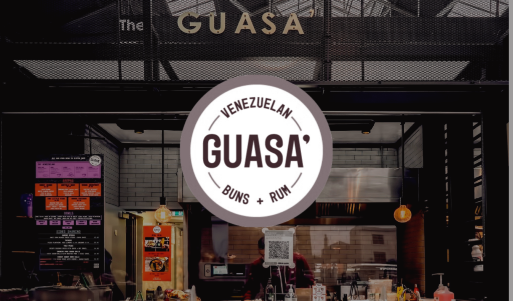 Front view of Guasa, a Venezuelan food stall with a circular logo reading "GUASA Venezuelan Buns Rum," located in a modern market setting.