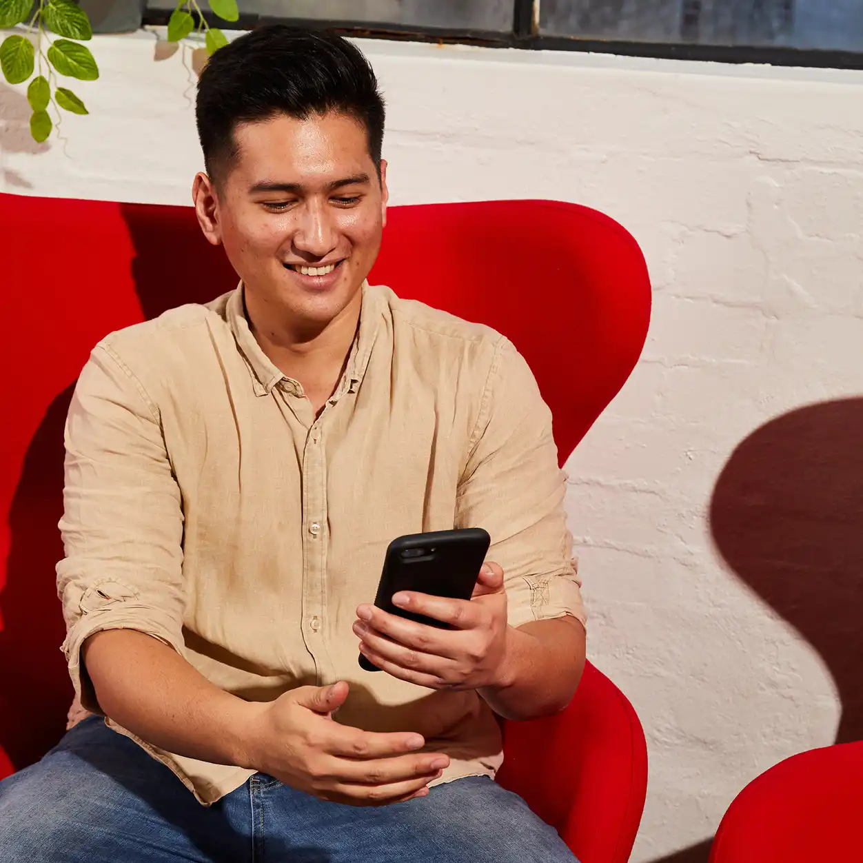 A man in a beige shirt smiles while looking at his phone, sitting in a vibrant red chair. Sunlight casts a shadow on the white brick wall behind.