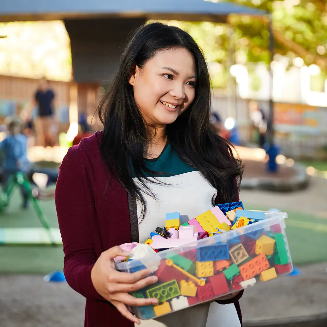 A woman smiles while holding a large plastic container filled with colorful building blocks. She stands in a sunny outdoor playground setting.