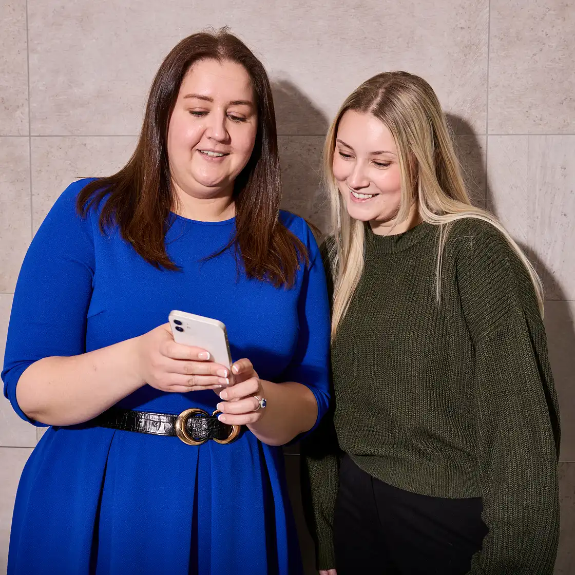 Two women standing together, the one on the left in a blue dress showing something on a phone to the other in a green sweater, both smiling warmly.