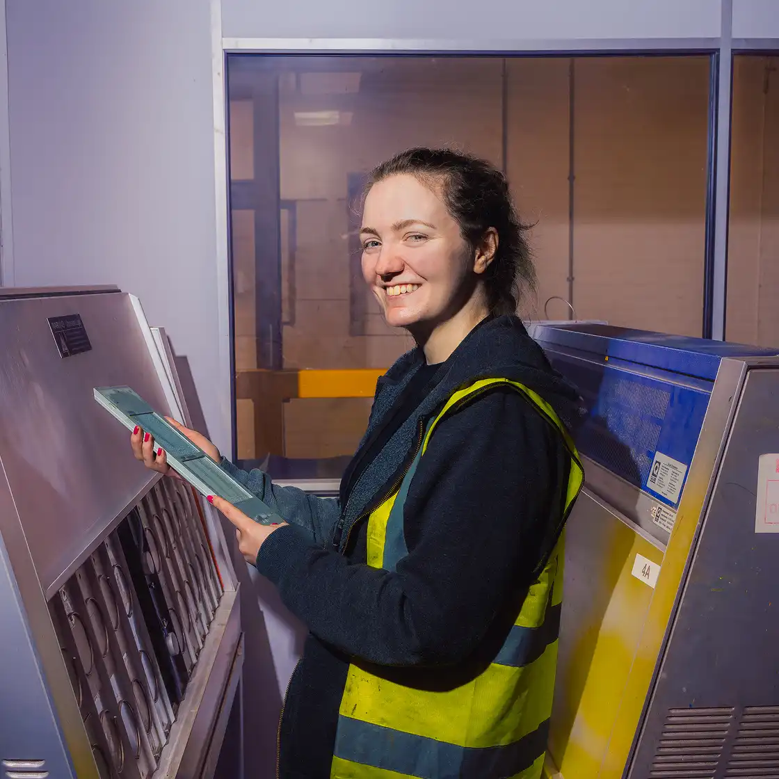 Smiling woman in a yellow safety vest stands at industrial machinery, holding a clipboard. The setting conveys a professional, cheerful atmosphere.
