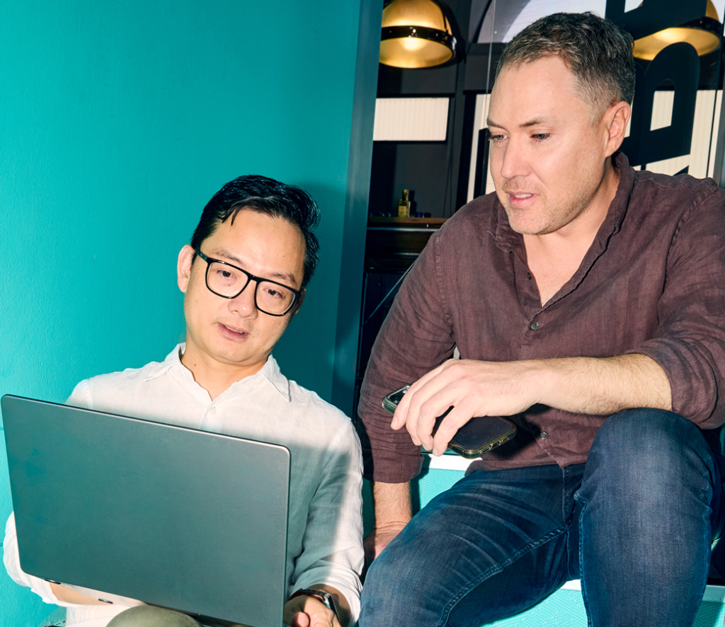 Two men sit together, one holding a laptop and the other a phone, engaged in a focused discussion against a teal wall background.
