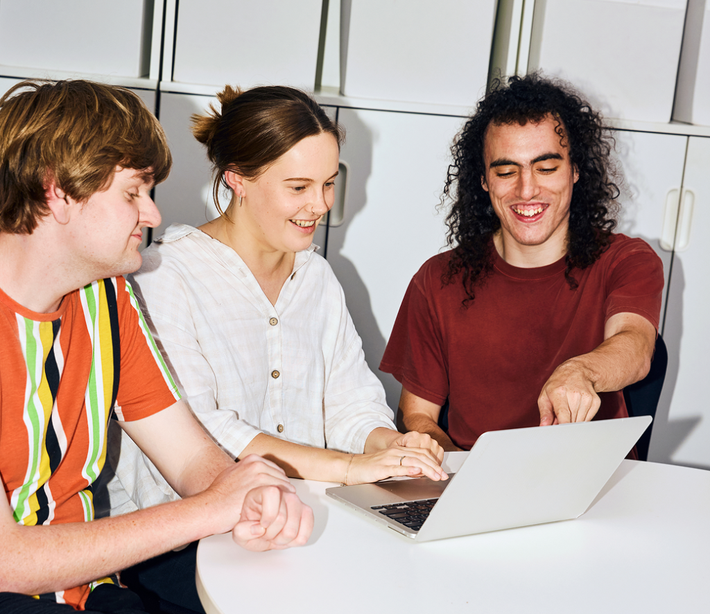 Three smiling young adults gathered around a laptop at a round table.