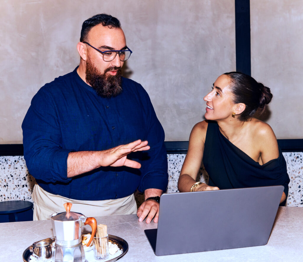 A man and a woman having a discussion at a counter while looking at a laptop; the man gestures with his hand, and a coffee set is placed in front of them.