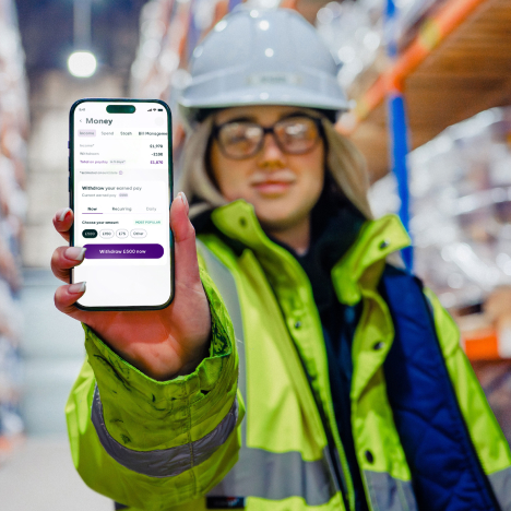 A woman in a hard hat and high-visibility jacket holds up a smartphone displaying a "Money" app, blurred warehouse shelves in the background.