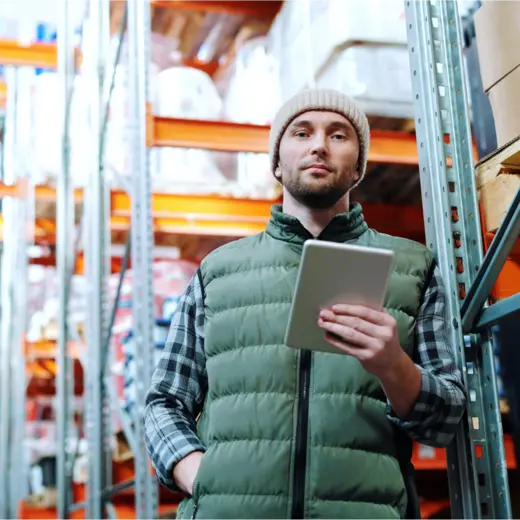 A man in a beanie and green quilted vest holds a tablet while standing in a warehouse aisle, surrounded by metal shelving stocked with various goods. He appears to be conducting inventory or managing stock.