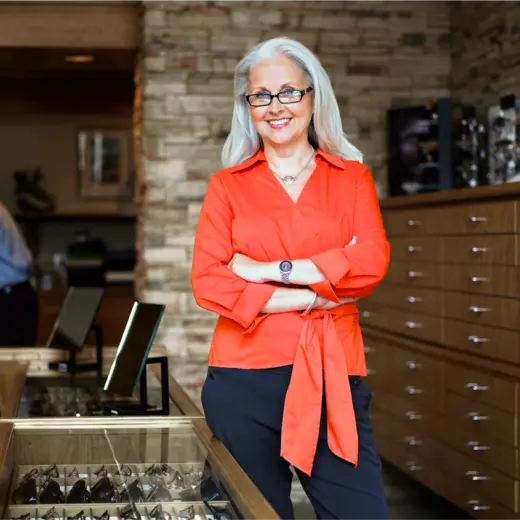 A confident older woman with long gray hair and glasses, wearing a bright orange blouse and black pants, stands with arms crossed in a stylish eyewear store. She is smiling and surrounded by display cases filled with glasses and sunglasses.