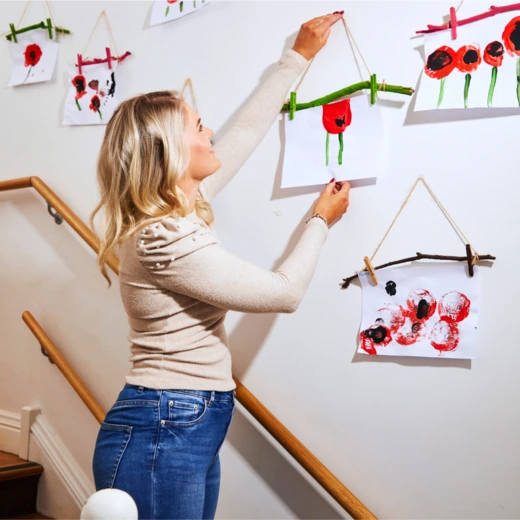 A woman with blonde hair wearing a beige sweater and blue jeans hangs children's artwork made with paint and sticks on a wall along a staircase. The artwork features colorful flower-like designs in red, green, and black.
