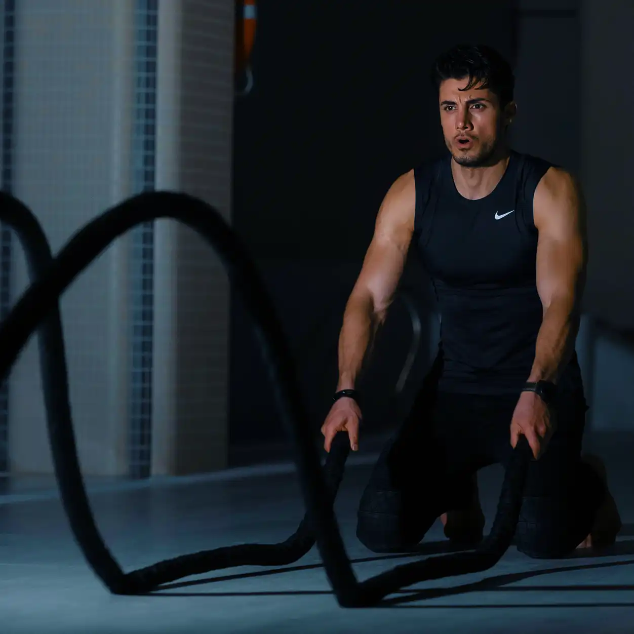 Man kneeling while exercising with battle ropes in a gym.